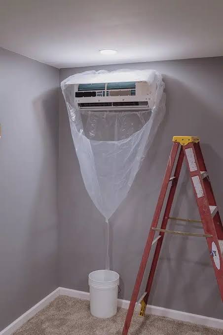 A bib bag taped around a wall-mounted mini-split, funnelling wash water into a bucket on the floor